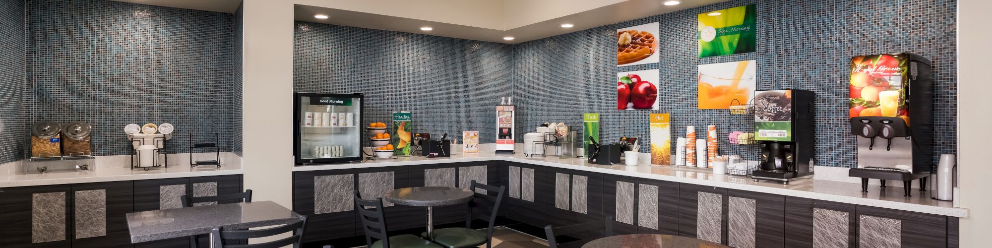A brightly lit self-serve breakfast area in a hotel with a variety of food and drink options, including a cereal dispenser, coffee maker, and juice machine.