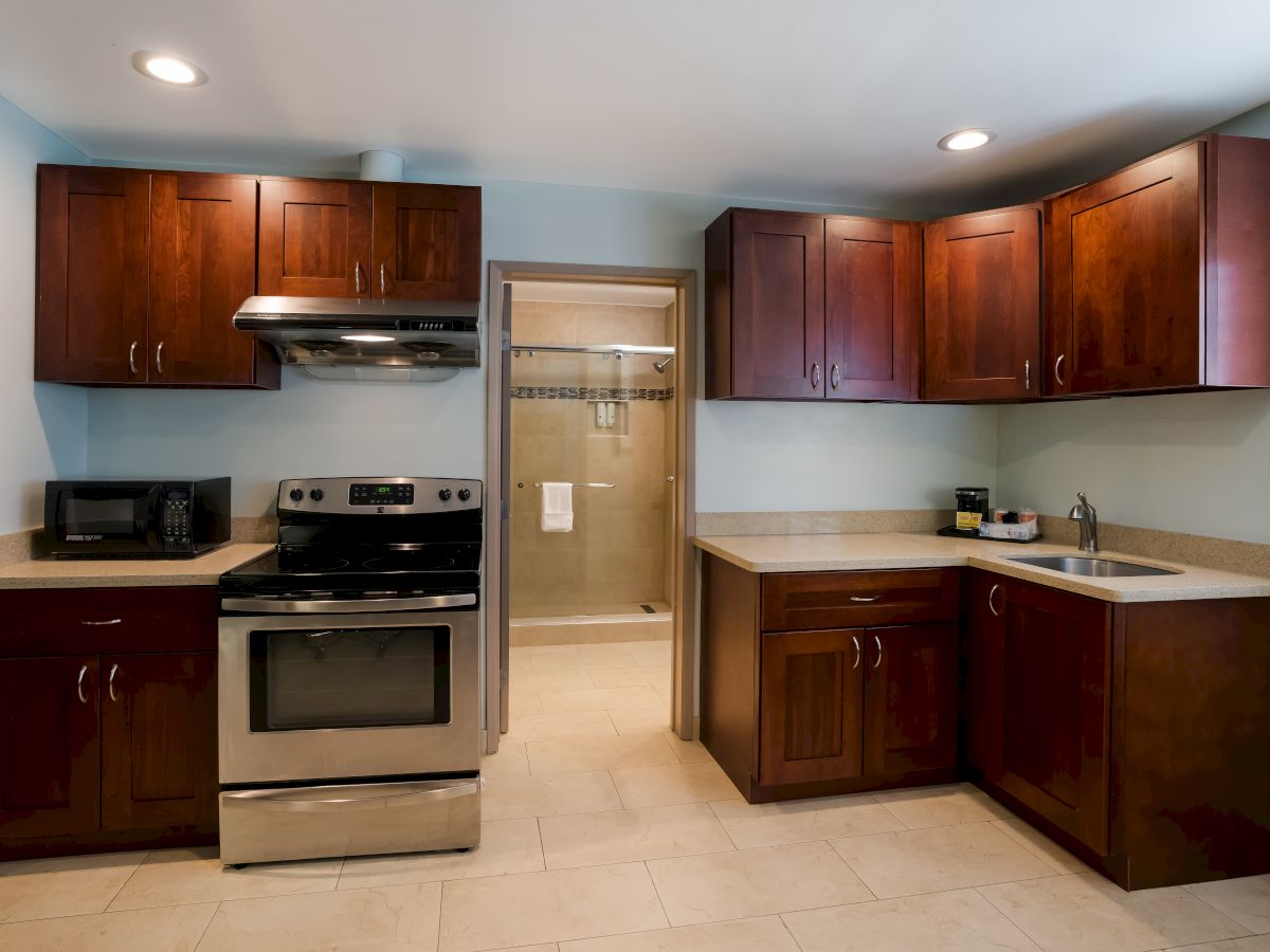 A modern kitchen with dark wooden cabinets, stainless steel appliances, a sink, and an adjoining bathroom in the background.