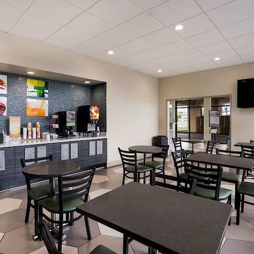 A clean, modern dining area with tables, chairs, beverage machines, and food displays; a TV is mounted on the wall.