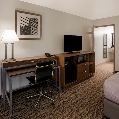 A hotel room with a bed, desk, chair, lamp, TV, and a small fridge. A doorway leads to a bathroom or another area of the room.