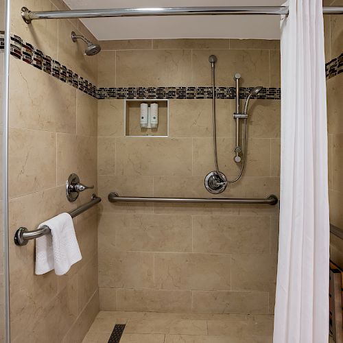 A spacious, accessible shower with grab bars, hand-held showerhead, soap dispensers, towel rack, and a curtain for privacy.