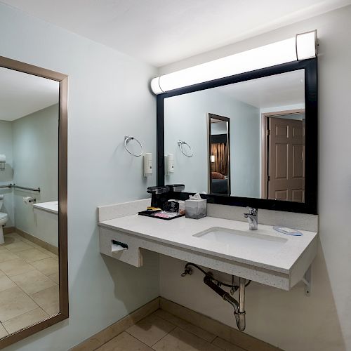 A bathroom featuring a sink with a large mirror, a wall-mounted hairdryer, toiletries, and a large framed mirror with the toilet area visible in the background.