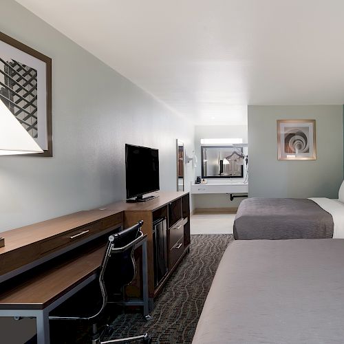 The image shows a hotel room with two beds, a TV, a work desk, and wall art. The room has modern decor and appears clean and organized.