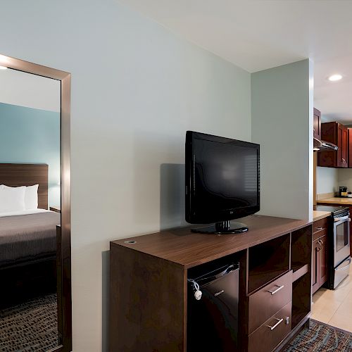 A modern hotel room features a bed, large mirror, flat-screen TV on a wooden cabinet, and a kitchenette with dark wood cabinets and stainless steel appliances.