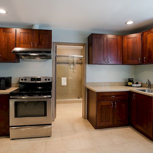 This image shows a kitchen with dark wood cabinets, stainless steel appliances, and beige countertops. There is a doorway leading to a bathroom.