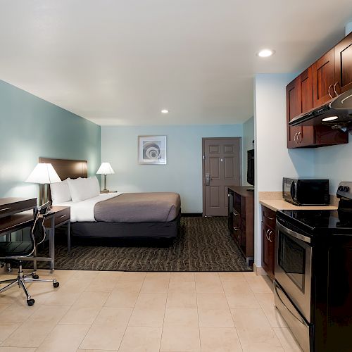 The image shows a hotel room with a bed, desk, chairs, and a small kitchen area with a stove, microwave, and cabinets. The walls are light blue.