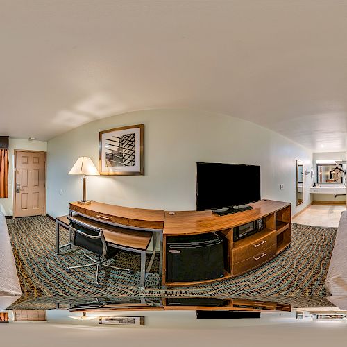This image shows a panoramic view of a hotel room with two beds, a TV on a wooden stand, a desk, and chairs. The room has modern decor and carpeting.